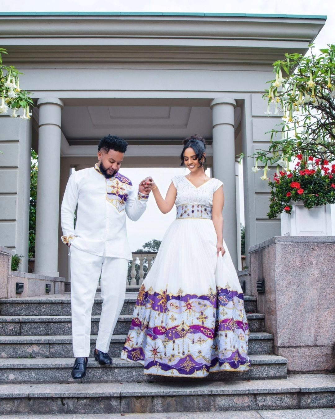 Couple in formal attire standing on steps outdoors with a building and flowers in the background. Festive Harmony Traditional Ethiopian Couples' Holiday Attire